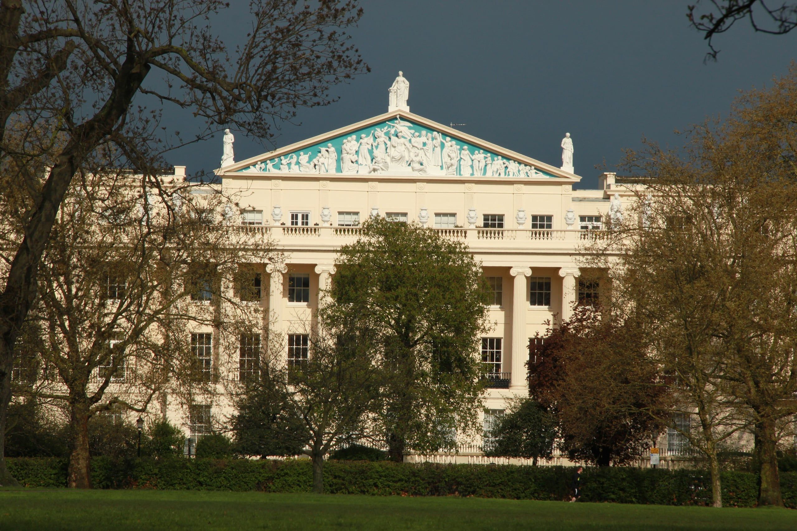 A white stucco Regency terrace with a sculpted pediment in Regent’s Park, London, lit by evening sun beneath a dark sky.